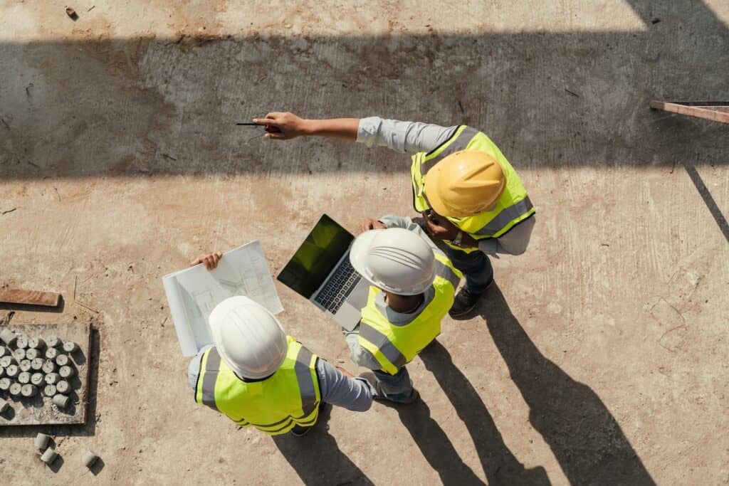 Construction workers discussing plans on site with laptop and blueprints.